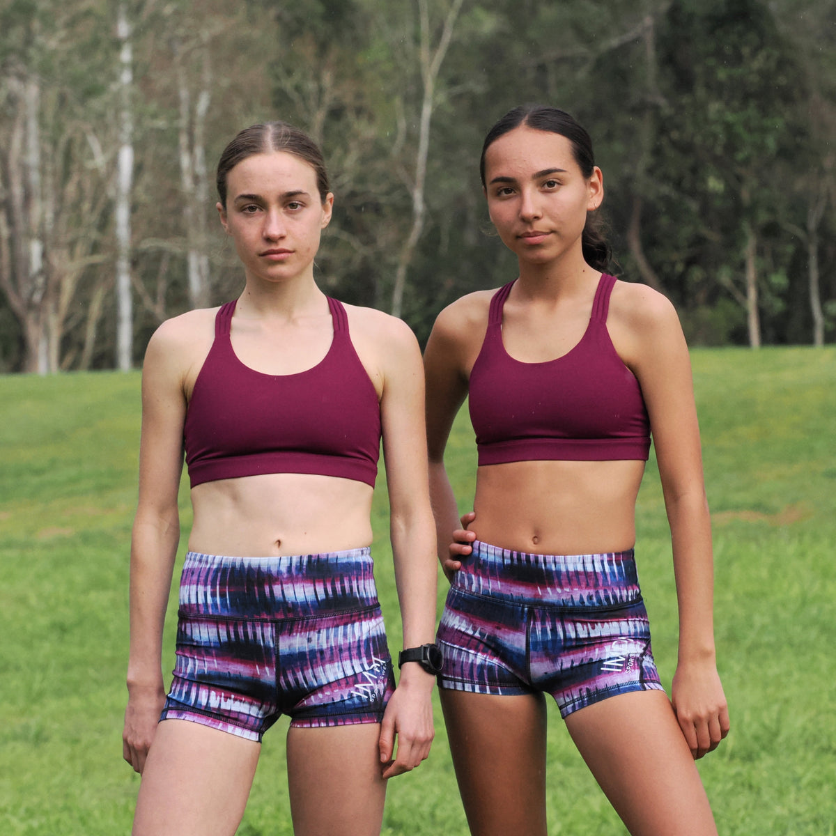 Two women in athletic wear standing outdoors on a grassy field with trees in the background.